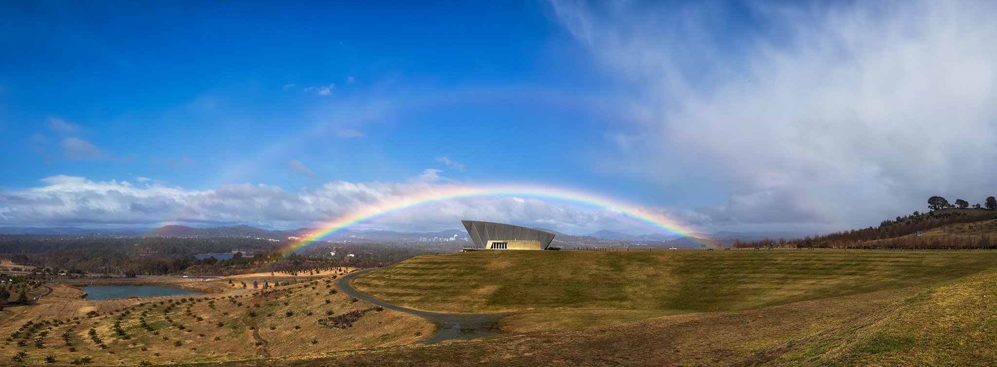 A full rainbow with a fainter second arc above it, framing the Margaret Whitlam Pavilion at the National Arboretum Canberra, photographed on a phone by Jacky Lee