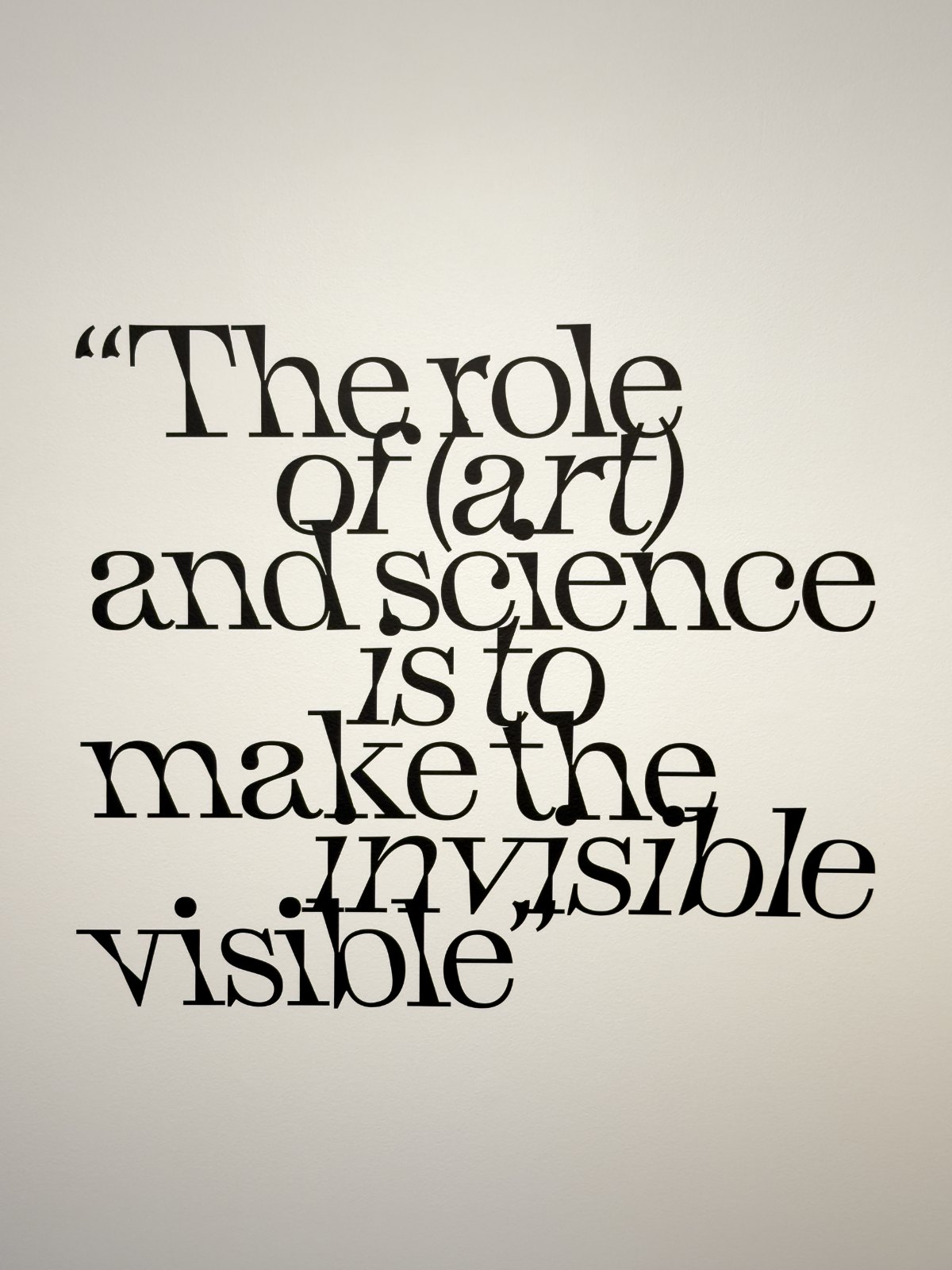 The quotation “The role of (art) and science is to make the invisible visible” in black lettering on the gallery wall