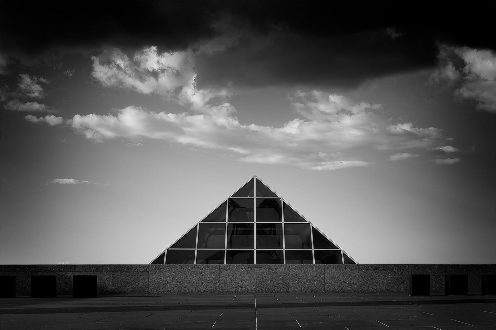 A glass pyramid rising behind a long stone wall under dark clouds, on Capital Hill, Canberra, in black and white