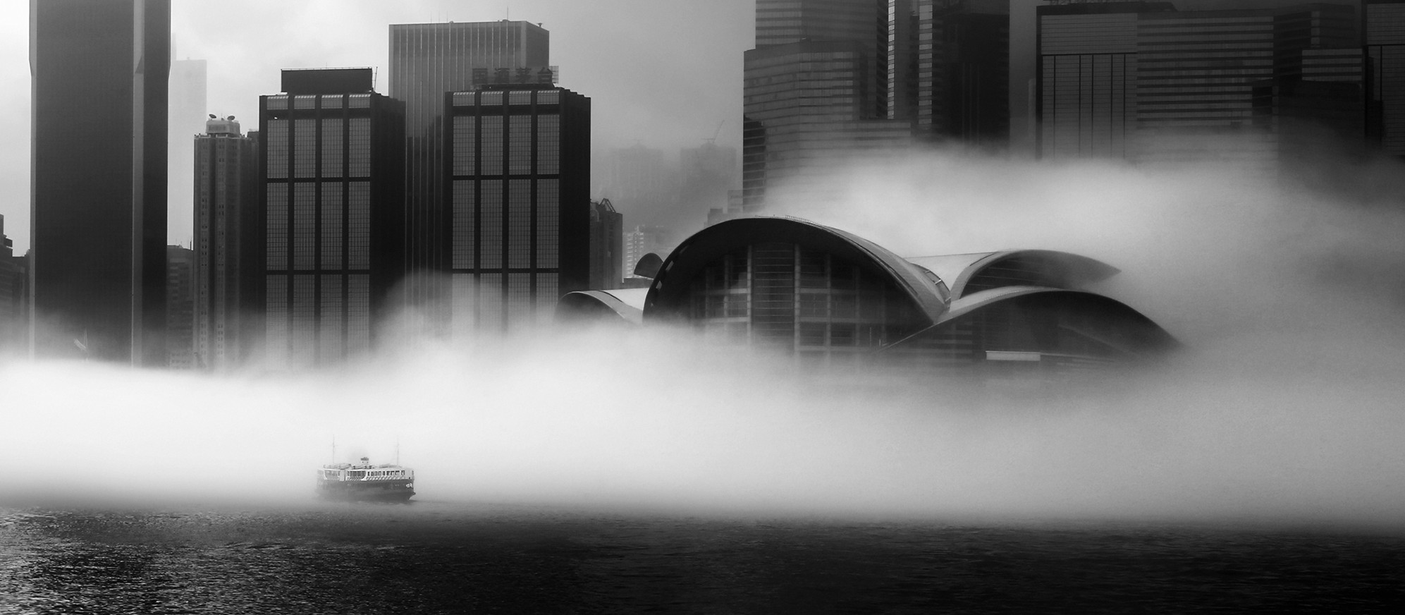 “A Foggy Morning”: a Star Ferry crosses fog-bound Victoria Harbour, Hong Kong, with the curved roof of the Hong Kong Convention and Exhibition Centre and tall towers rising from the mist behind it, in a black-and-white photograph by Jacky Lee